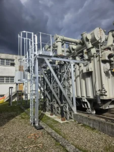 Travaux d'installation d'équipements de protection collective et individuelle sur un transformateur électrique haute tension par la sarl HTB Height Technician in Building ; systèmes antichute via échelle à crinoline avec passerelle avec garde-corps pour un accès sécurisé au transformateur, dans le cadre de travaux de sécurisation contre les chutes de hauteur