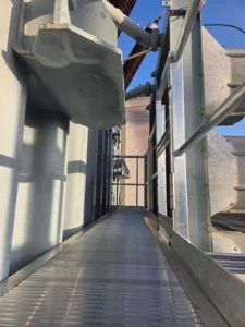 Intervention de Height Technician in Building sur un transformateur électrique pour la pose d'un équipement de protection collective. Il s'agit là d'installer une passerelle avec garde-corps dans le cadre de travaux de sécurisation contre les chutes de hauteur. La passerelle est munie d'un support d'échelle parallèle.