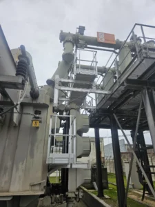 Intervention de Height Technician in Building sur un transformateur électrique pour la pose d'équipements de protection collective. Il s'agit là d'installer trois passerelles avec garde-corps dans le cadre de travaux de sécurisation contre les chutes de hauteur. Deux sont munies d'un support d'échelle parallèle, la dernière d'une échelle à crinoline.