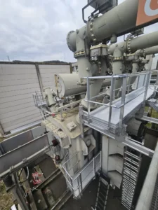 Intervention de Height Technician in Building sur un transformateur électrique pour la pose d'équipements de protection collective. Il s'agit là d'installer trois passerelles avec garde-corps dans le cadre de travaux de sécurisation contre les chutes de hauteur. Deux sont munies d'un support d'échelle parallèle, la dernière d'une échelle à crinoline.